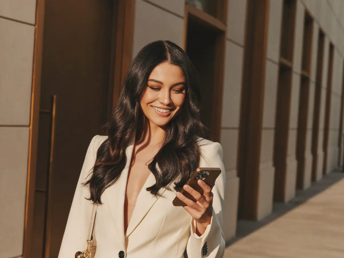 A professional in a cream blazer smiling at her phone as she walks along a sunlit street